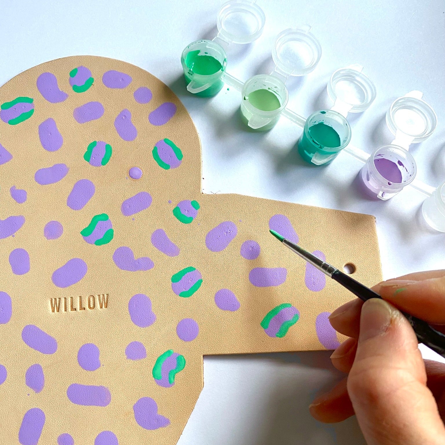 Person painting leopard pattern on leather purse cutout with purple and green paint on a white background.