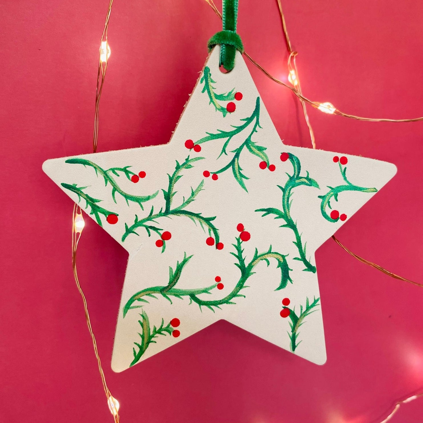 Star-shaped Christmas ornament with greenery and berries on a pink background with string lights.