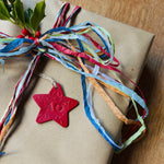 Load image into Gallery viewer, A gift-wrapped package in brown kraft paper, tied with multicolored paper ribbons and a red star-shaped ornament, placed on a wooden surface.
