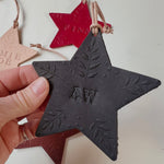 Load image into Gallery viewer, A hand holding a dark charcoal gray star-shaped ornament with leaf engravings and the monogram 'AW' in the center. Other star ornaments in red and beige are partially visible in the background against a plain off-white surface.
