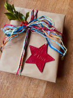 Load image into Gallery viewer, A wrapped gift box on a wooden surface, wrapped in brown paper with a multicolored ribbon bow and a red star ornament embossed with the name 'FINN'.
