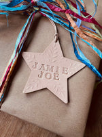 Load image into Gallery viewer, A personalized star-shaped wooden ornament engraved with 'JAMIE JOE' and tied to a brown kraft paper gift box with colorful paper streamers.
