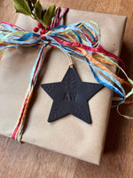 Load image into Gallery viewer, A black star-shaped monogrammed ornament with leaf engravings hangs from a kraft paper gift box tied with colorful paper ribbons and a sprig of holly on a wooden surface.
