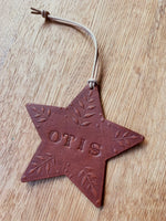 Load image into Gallery viewer, A brown leather star-shaped ornament with the name 'OTIS' embossed in the center and leaf designs around it, hanging from a light tan cord on a wooden surface.

