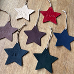 Load image into Gallery viewer, Six embossed star-shaped leather ornaments in different colors (cream, red, mauve, dark purple, navy, and teal) arranged on a light wood background. Each ornament has a hanging loop and is personalized with an engraved name or initials.
