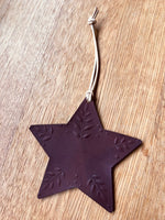 Load image into Gallery viewer, A dark brown star-shaped ornament with embossed leaf patterns, hanging from a light-colored cord on a wooden surface.
