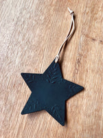 Load image into Gallery viewer, A black star-shaped hanging ornament with embossed leaf details and a natural twine loop, displayed on a light brown wooden surface.
