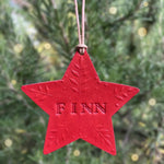 Load image into Gallery viewer, A red star-shaped hanging ornament with the name 'FINN' embossed in the center, suspended by a thin string against a blurred green outdoor background with soft yellow bokeh lights.
