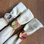 Load image into Gallery viewer, Three cream-colored textured leather  napkins tied with circular paper napkin rings featuring marbled patterns in blue, green, pink, and red, arranged on a brown wooden surface.

