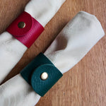 Load image into Gallery viewer, Two rolled white napkins on a wooden surface, each secured with a leather napkin holder strap in red and dark teal, both with metal snap buttons.
