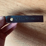 Load image into Gallery viewer, Hand holding a leather keychain with &#39;GET HOME SAFE X&#39; text on a wooden surface
